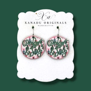May include: A pair of round dangle earrings featuring a pink and green Christmas tree pattern with the dark green text Comfort & Joy layered on top. The earrings are displayed on a white card labeled Xo Xanadu Originals Handcrafted.