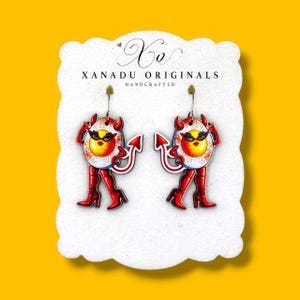 May include: A pair of dangle earrings featuring a cartoon egg character with devil horns, a tail, and red high-heeled boots. The earrings are displayed on a white scalloped card labeled Xo XANADU ORIGINALS HANDCRAFTED. The design includes red and yellow colors.