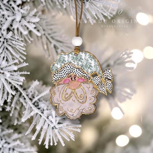 May include: A Santa Claus ornament featuring a light green hat with a white floral pattern, a black and white polka dot brim, and a matching bow. The figure has a pale pink face, a white beard with gold outlines, and hangs from a brown cord.