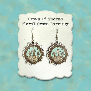 May include: Floral cross earrings with a crown of thorns design. The earrings feature three crosses on a light blue background, surrounded by a brown crown of thorns. The text "Crown Of Thorns Floral Cross Earrings" is displayed on the white card.