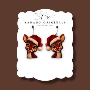 May include: A pair of dangle earrings featuring a brown reindeer head with a red nose and a red Santa hat with a white trim. The earrings are displayed on a white card with the text "XO XANADU ORIGINALS HANDCRAFTED" printed at the top.