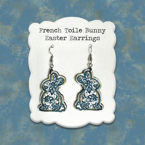 May include: A pair of French Toile Bunny Easter Earrings. The earrings are shaped like bunnies with a blue and white floral design. The earrings are displayed on a white, scalloped card against a blue background.