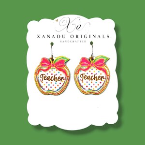 May include: A pair of dangle earrings shaped like apples with a white background, colorful polka dots, and a bright pink bow. The word Teacher is written in brown script across the center of each apple. The earrings are displayed on a white card.