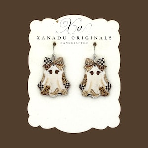 May include: A pair of ghost-shaped dangle earrings featuring a cream-colored body with brown leopard print and black and white checkered patterns. Each ghost has a bow on top and is displayed on a white card labeled Xanadu Originals Handcrafted.