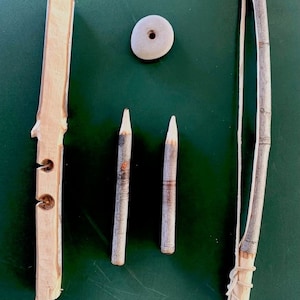 May include: A set of wooden tools for making fire. The set includes a fire drill, a hand drill, a fire board, a spindle, and a fire stone. The tools are laid out on a green surface.