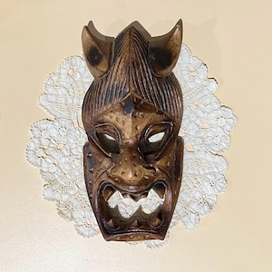 May include: A wooden mask with a carved face and horns. The mask is brown and has a detailed design.