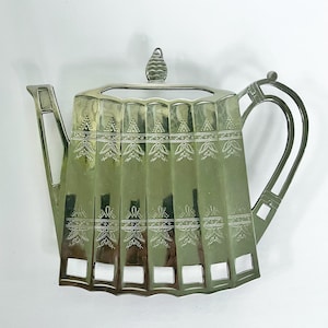 Puede incluir: Decoración de pared en forma de tetera de plata con un diseño decorativo. La tetera tiene un cuerpo acanalado y un asa detallada.