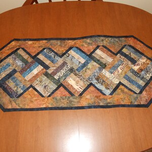 Peut inclure: Un chemin de table avec un motif g&eacute;om&eacute;trique dans des tons de brun, bleu et beige. Le chemin de table est fait de tissu avec un motif batik et a une bordure bleu fonc&eacute;.
