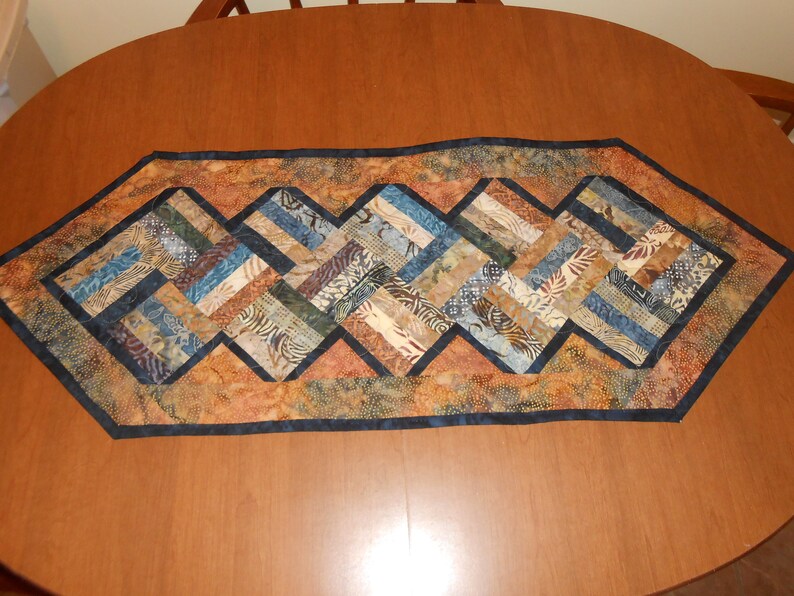 Peut inclure: Un chemin de table avec un motif g&eacute;om&eacute;trique dans des tons de brun, bleu et beige. Le chemin de table est fait de tissu avec un motif batik et a une bordure bleu fonc&eacute;.