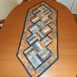 Peut inclure: Un chemin de table matelass&eacute; avec un motif g&eacute;om&eacute;trique dans des tons de brun, bleu et beige. Le chemin de table a une bordure bleu fonc&eacute; et est fait de tissu patchwork.