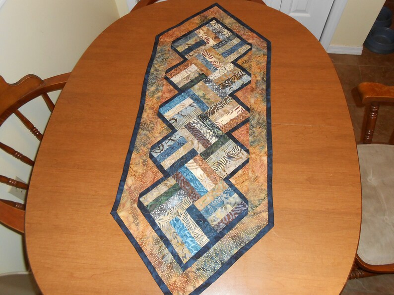 Peut inclure: Un chemin de table matelass&eacute; avec un motif g&eacute;om&eacute;trique dans des tons de brun, bleu et beige. Le chemin de table a une bordure bleu fonc&eacute; et est fait de tissu patchwork.