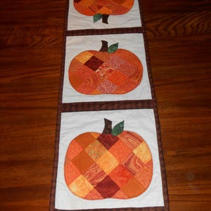 May include: A white table runner with three patchwork pumpkin designs. The pumpkins are made of various shades of orange, brown, and red fabric.