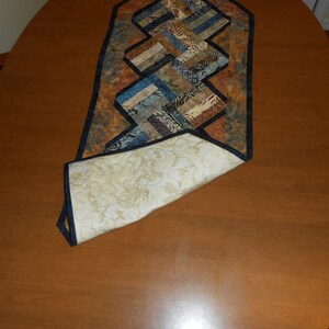 Peut inclure: Un chemin de table matelass&eacute; avec un motif g&eacute;om&eacute;trique dans des tons de brun, bleu et beige. Le chemin de table a une bordure noire et est pli&eacute; en deux, r&eacute;v&eacute;lant un dos matelass&eacute; blanc.