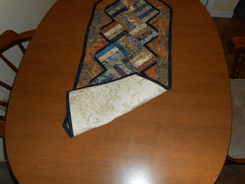 Peut inclure: Un chemin de table matelass&eacute; avec un motif g&eacute;om&eacute;trique dans des tons de brun, bleu et beige. Le chemin de table a une bordure noire et est pli&eacute; en deux, r&eacute;v&eacute;lant un dos matelass&eacute; blanc.