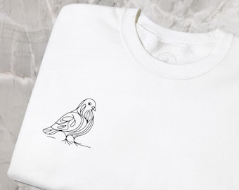 May include: White crew neck sweatshirt with a minimalist black line drawing of a bird on the left chest. The bird is perched on a thin line, with detailed feather and wing outlines. The sweatshirt is folded neatly on a marble surface.