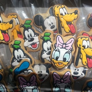 May include: A collection of colorful, decorated sugar cookies featuring various Disney characters, including Mickey Mouse, Minnie Mouse, Goofy, Donald Duck, and Pluto. The cookies are arranged in a clear plastic bag.