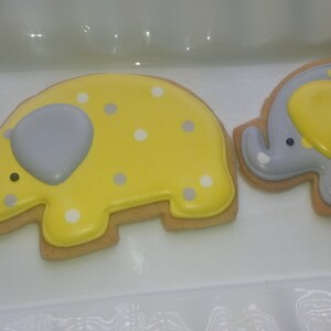 May include: Two decorated sugar cookies shaped like elephants. One elephant is yellow with white polka dots and a gray trunk. The other elephant is gray with white polka dots and a yellow trunk.