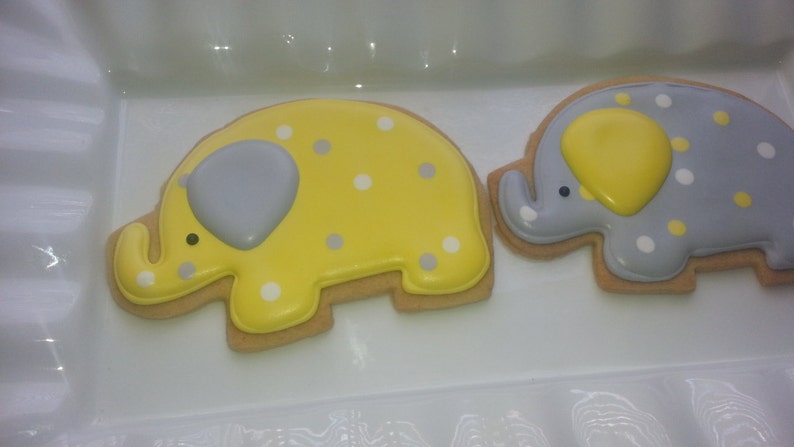 May include: Two decorated sugar cookies shaped like elephants. One elephant is yellow with white polka dots and a gray trunk. The other elephant is gray with white polka dots and a yellow trunk.