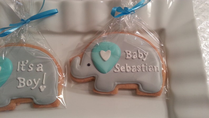 May include: Two decorated sugar cookies shaped like elephants. One cookie is light blue with the text "It's a Boy!" and a blue heart. The other cookie is light blue with the text "Baby Sebastian" and a blue heart.
