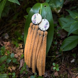 May include: A pair of dangle earrings featuring circular off-white wooden discs with a small floral print of purple and green sprigs. Each disc is attached to multiple long, tan-coloured suede leather fringe strips hanging from silver-toned metal hooks.
