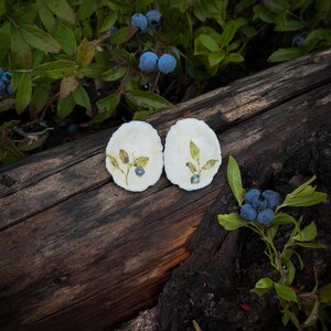 May include: Two oval-shaped white ceramic dishes featuring a botanical illustration of a green leaf sprig and a single blue blueberry. The dishes have an irregular, scalloped edge and a smooth, matte finish, displayed as a pair on a dark wooden surface.