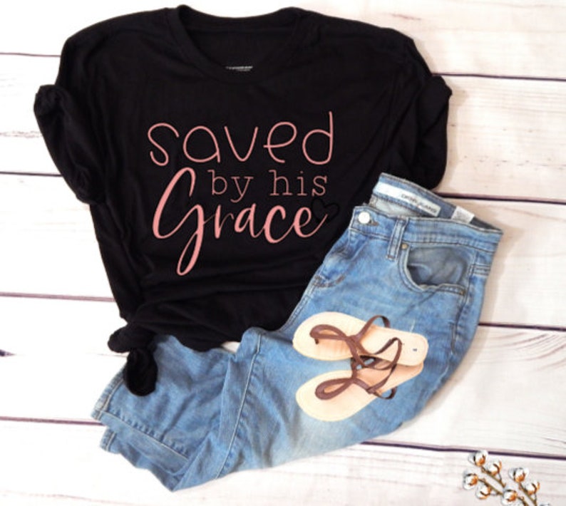 K&ouml;nnte beinhalten: Schwarzes T-Shirt mit dem Text "saved by his grace" in rosa Schrift. Das Shirt wird mit einer blauen Jeans und braunen Sandalen kombiniert.