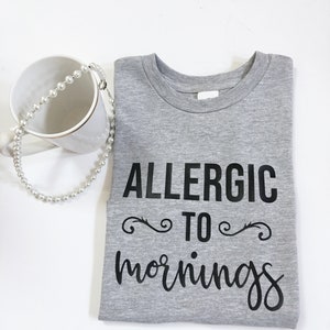 May include: A gray t-shirt with the words "ALLERGIC TO mornings" in black script. A white mug and a pearl necklace are next to the shirt.