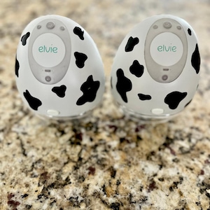 May include: Two white and black cow print electric breast pumps with the word "elvie" on the front.