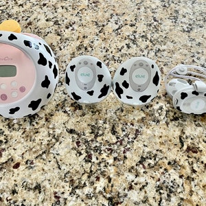 May include: A pink and white electric breast pump with a digital display and a cow print design. Two white and black cow print wireless breast pump attachments and a pair of white and black cow print headphones are also shown.