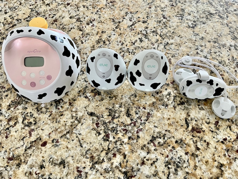 May include: A pink and white electric breast pump with a digital display and a cow print design. Two white and black cow print wireless breast pump attachments and a pair of white and black cow print headphones are also shown.