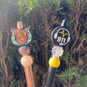 May include: Two pens with decorative toppers. One features a Highland cow design with floral accents, the other a 911 emergency services theme with headphones. Both pens have beaded accents and metallic or black barrels.