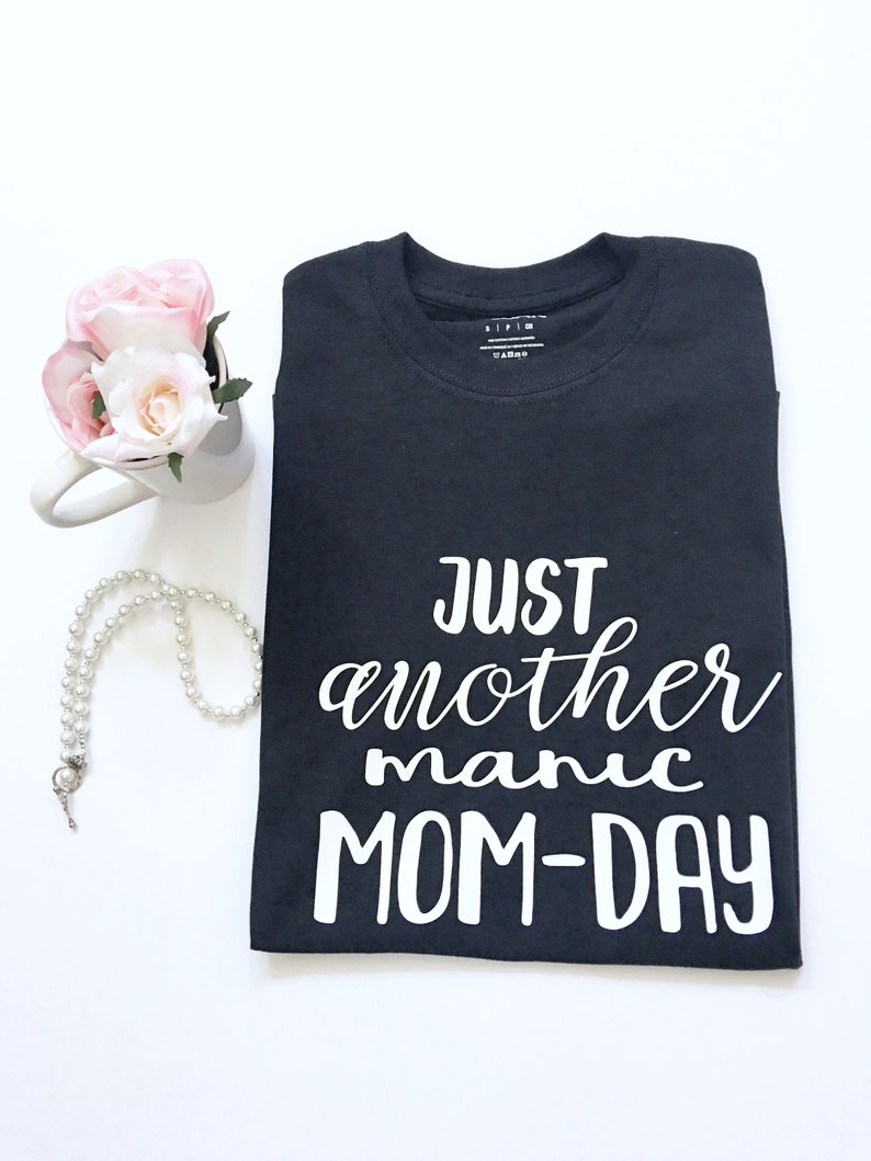 May include: Black t-shirt with white text that reads "Just another manic MOM-DAY".