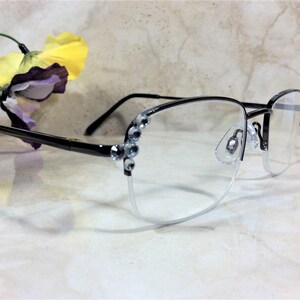 May include: Semi-rimless eyeglasses with a black frame and clear lenses. The upper frame is black, and the lower frame is a silver color. The temples are black, and the outer corners of the frame are decorated with clear rhinestones. A yellow and purple flower is in the background.