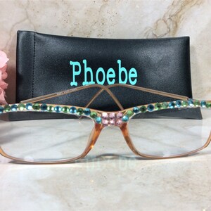 May include: Eyeglasses with a decorative design. The frames are a light brown color, with clear lenses. The top of the frames are adorned with pink, green, and blue rhinestones. A black case with the name "Phoebe" in teal is behind the glasses.