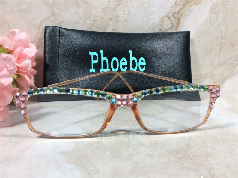 May include: Eyeglasses with a decorative design. The frames are a light brown color, with clear lenses. The top of the frames are adorned with pink, green, and blue rhinestones. A black case with the name "Phoebe" in teal is behind the glasses.