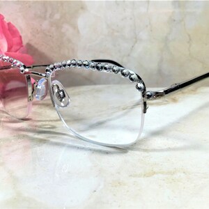 May include: Silver-framed eyeglasses with clear lenses. The top of the frames are embellished with a row of sparkling rhinestones. The temples are black. The glasses are set against a marble background with a pink flower in the background.