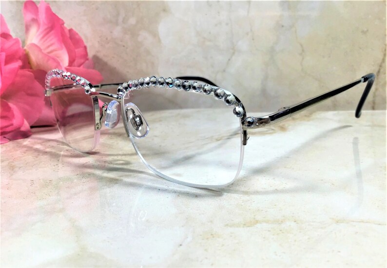 May include: Silver-framed eyeglasses with clear lenses. The top of the frames are embellished with a row of sparkling rhinestones. The temples are black. The glasses are set against a marble background with a pink flower in the background.