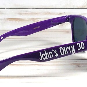 K&ouml;nnte beinhalten: Violette Sonnenbrille mit dunklen Gl&auml;sern. Der Text "John's Dirty 30" ist wei&szlig; auf dem linken B&uuml;gel aufgedruckt. Die Sonnenbrille ist abgewinkelt und zeigt die Seite und die Vorderseite.