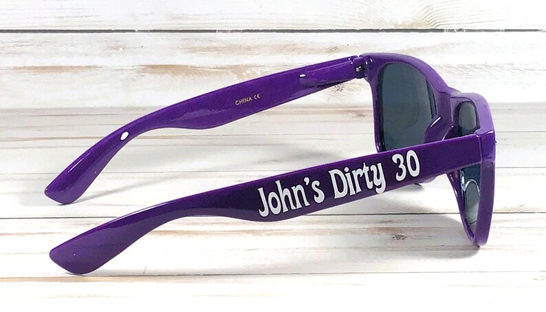 K&ouml;nnte beinhalten: Violette Sonnenbrille mit dunklen Gl&auml;sern. Der Text "John's Dirty 30" ist wei&szlig; auf dem linken B&uuml;gel aufgedruckt. Die Sonnenbrille ist abgewinkelt und zeigt die Seite und die Vorderseite.