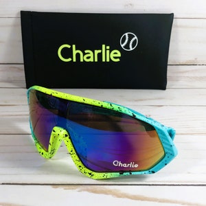 Puede incluir: Un par de gafas de sol azules, verdes y amarillas con un dise&ntilde;o de pintura negra salpicada. Las gafas de sol tienen lentes espejadas y dicen "Charlie" en el marco.