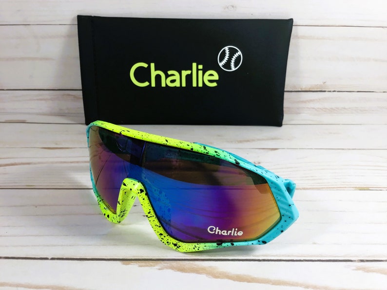 Puede incluir: Un par de gafas de sol azules, verdes y amarillas con un dise&ntilde;o de pintura negra salpicada. Las gafas de sol tienen lentes espejadas y dicen "Charlie" en el marco.