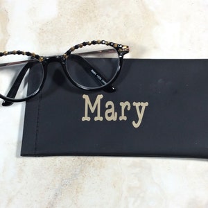 May include: Round-framed black spectacles with decorative detailing on the upper rims sit on a black glasses case. The case is personalised with the name "Mary" in gold lettering.