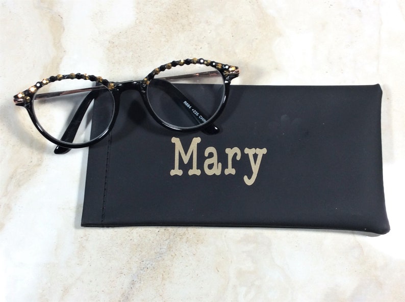 May include: Round-framed black spectacles with decorative detailing on the upper rims sit on a black glasses case. The case is personalised with the name "Mary" in gold lettering.