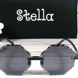 May include: Octagon shaped sunglasses with a black frame and dark gray lenses. The sunglasses are on a white wooden surface. The word "Stella" is printed on a black surface behind the sunglasses.