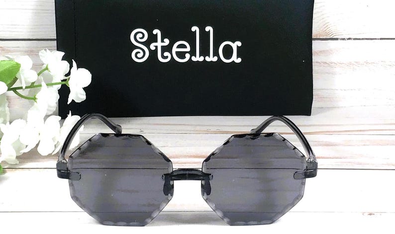 May include: Octagon shaped sunglasses with a black frame and dark gray lenses. The sunglasses are on a white wooden surface. The word "Stella" is printed on a black surface behind the sunglasses.