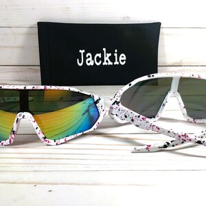 Puede incluir: Gafas de sol blancas con un dise&ntilde;o de pintura salpicada en negro y rosa. Las gafas de sol tienen lentes espejadas y vienen con una funda negra que dice "Jackie".