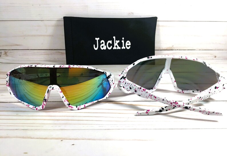 Puede incluir: Gafas de sol blancas con un dise&ntilde;o de pintura salpicada en negro y rosa. Las gafas de sol tienen lentes espejadas y vienen con una funda negra que dice "Jackie".