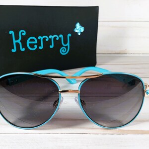 Pu&ograve; includere: Occhiali da sole aviator blu con dettagli dorati e strass. Gli occhiali hanno lenti scure e una montatura blu.