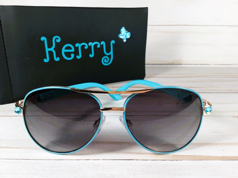 Pu&ograve; includere: Occhiali da sole aviator blu con dettagli dorati e strass. Gli occhiali hanno lenti scure e una montatura blu.