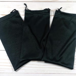 May include: Three black drawstring pouches are arranged on a light wood-grain surface. Each pouch is made of a smooth, dark fabric and has a drawstring closure at the top. The pouches are empty and ready for use.